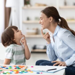 Speech training for kids. Professional woman specialist training with little boy at cabinet, teaching him right articulation exercises, side view